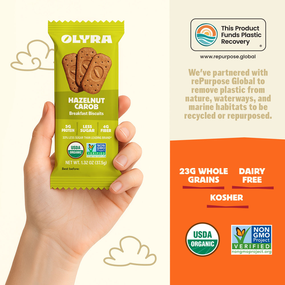 Hand holding a green Olyra hazelnut carob breakfast biscuit package with product details on an orange background.