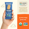 Hand holding a blue Olyra Greek Yogurt Blueberry breakfast biscuit package with product details on an orange background.