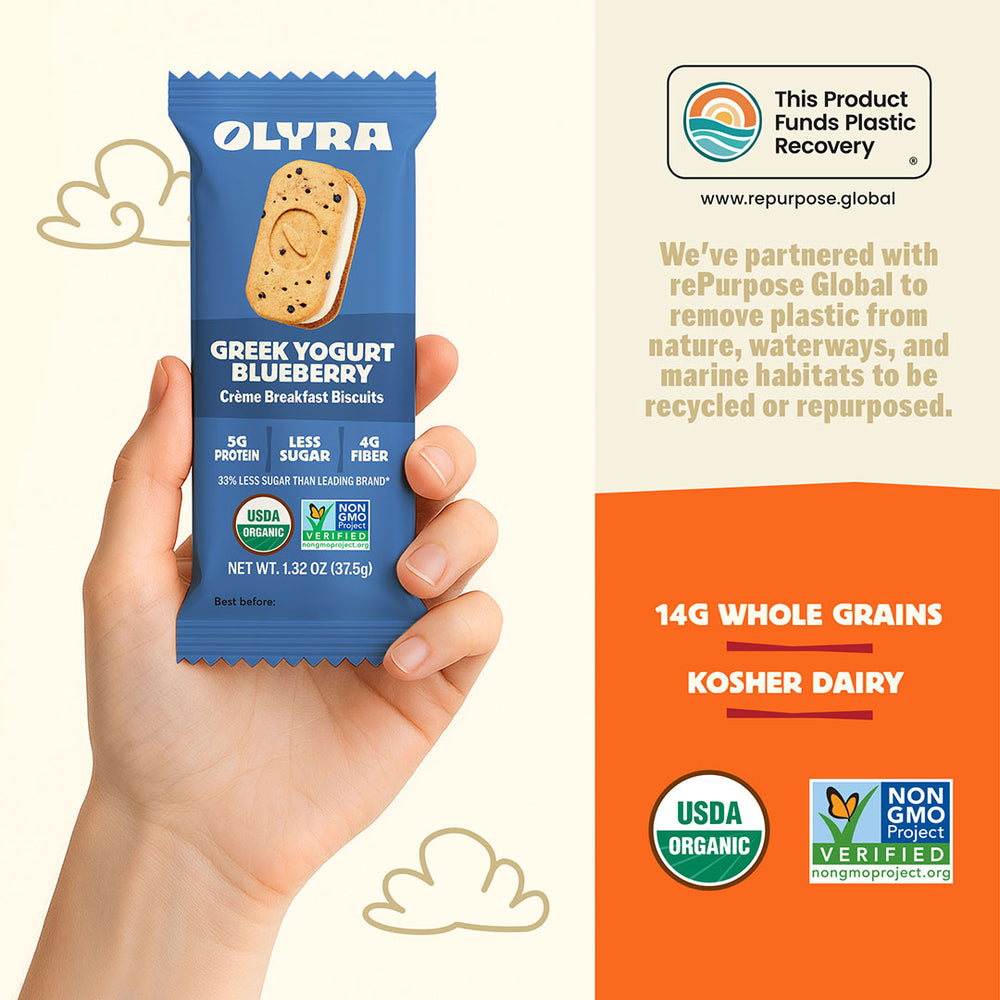 Hand holding a blue Olyra Greek Yogurt Blueberry breakfast biscuit package with product details on an orange background.