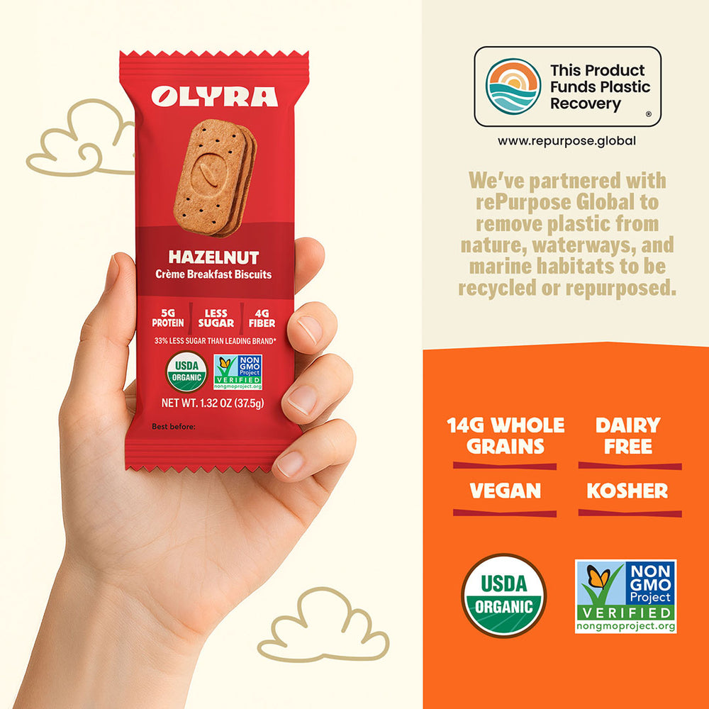 Hand holding a red Olyra hazelnut breakfast biscuit package with product details on an orange background.
