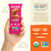 Olyra strawberry fruit and grain bar packaging held by a hand, with product details and certification logos on an orange background.