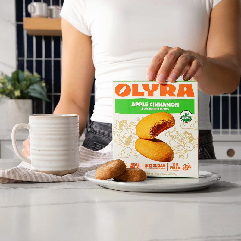 Person holding a box of Olyra Apple Cinnamon soft baked bites with cookies and a mug on a plate.