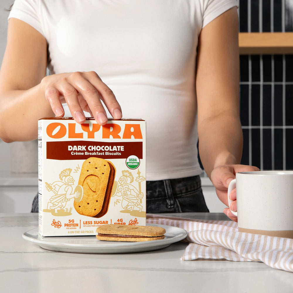 Person holding a box of Olyra dark chocolate creme breakfast biscuits with a biscuit sandwich on a plate.