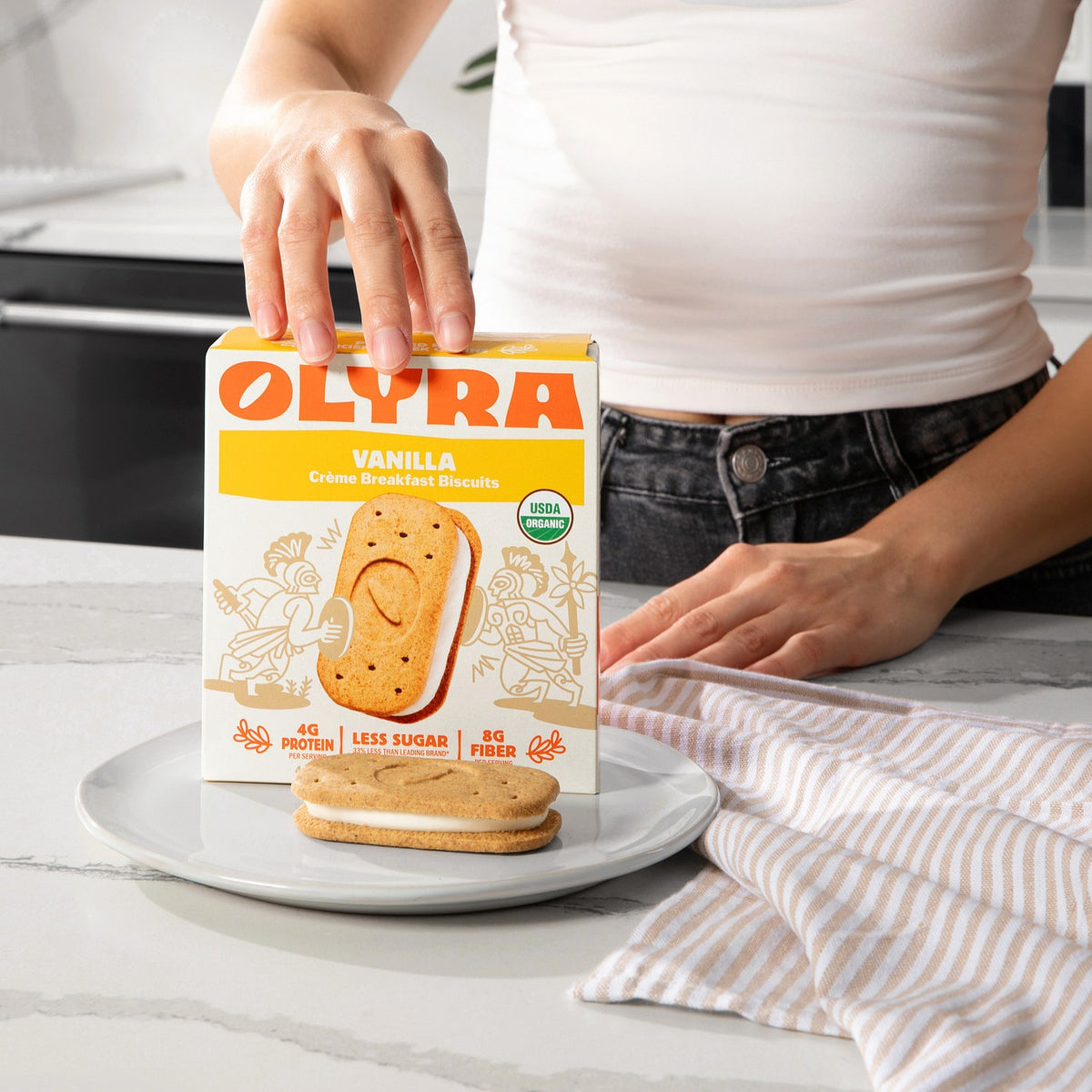 Person holding an Olyra vanilla creme sandwich box with a biscuit sandwich on a plate.