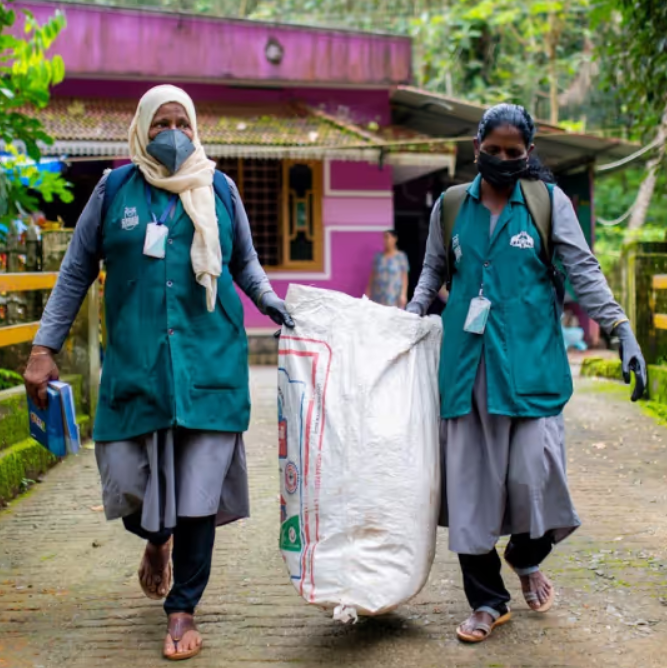 Two individuals in green uniforms carrying a large bag on a path with a building and trees in the background.