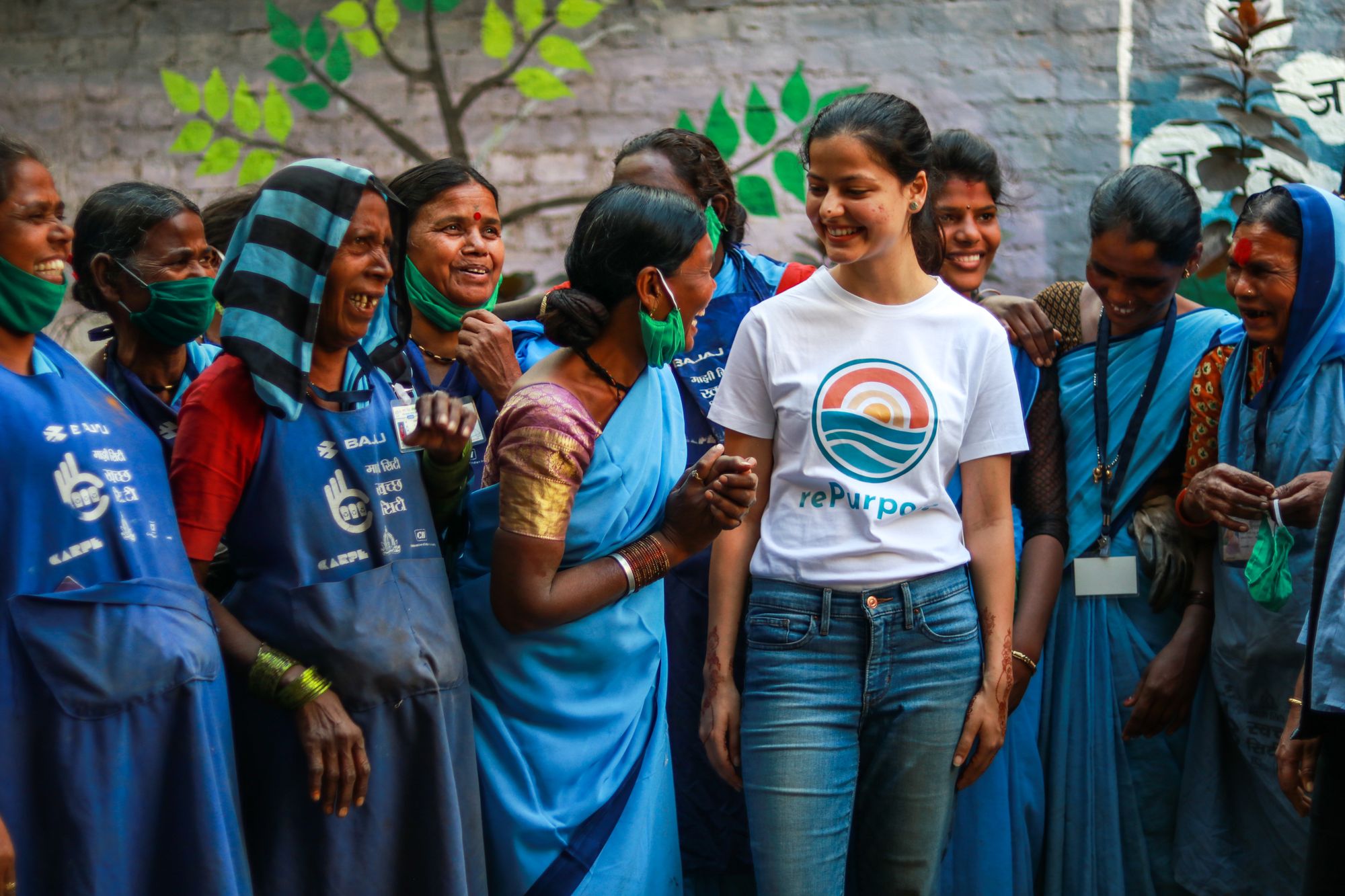 Woman in a white repurpose shirt standing among people wearing blue uniforms, with a mural in the background.