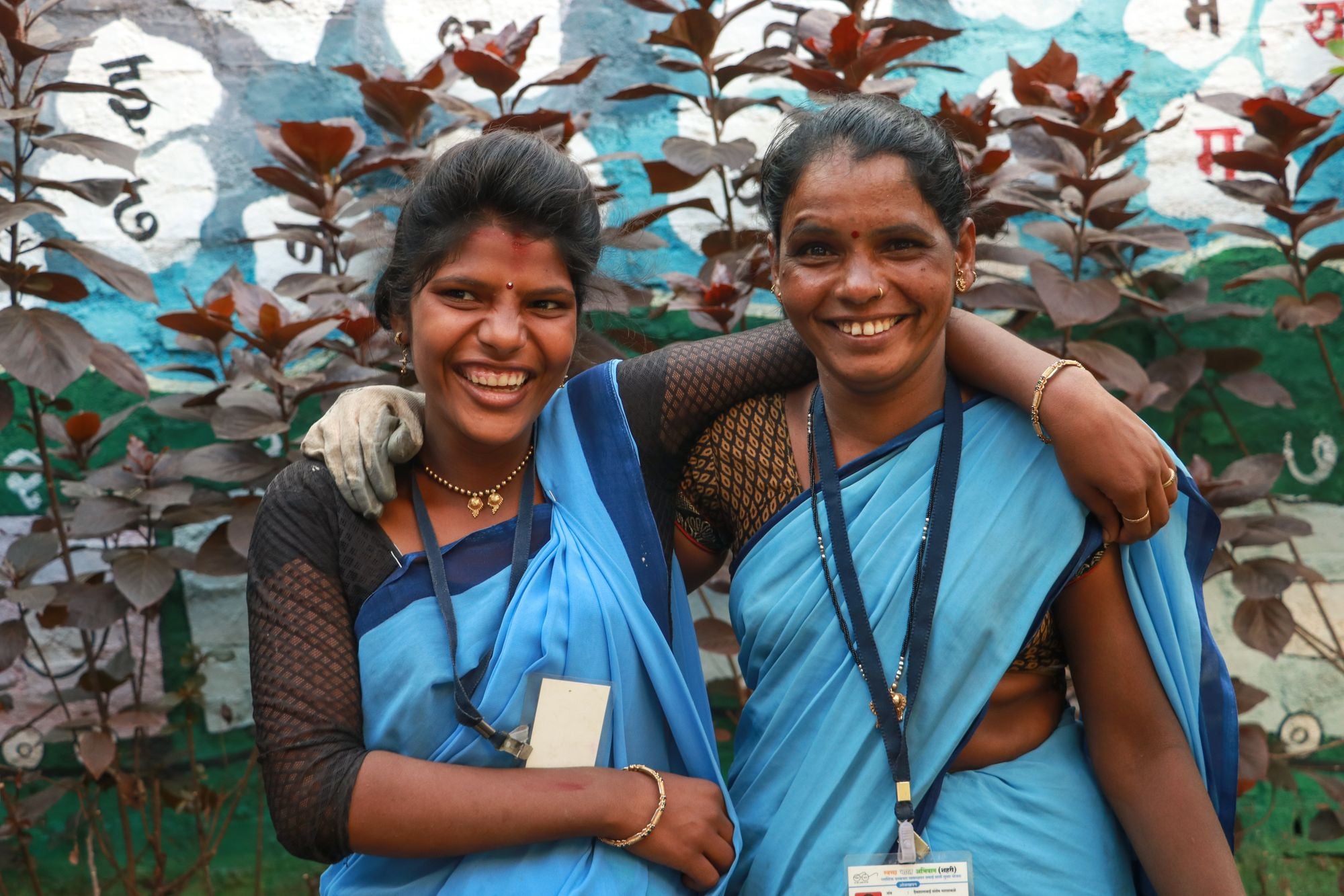 Two women in blue sarees standing in front of a colorful mural.